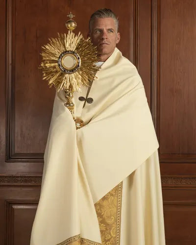 Man in religious attire holding a golden monstrance against a wooden paneled background