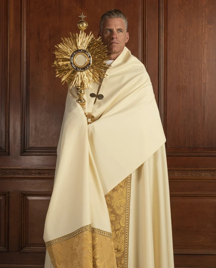 Priest in white robe holding a monstrance against a wooden paneled background